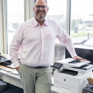 Justdata ICT, Een lachende man met bril en lichtroze overhemd staat naast een witte HP printer in een licht kantoor met grote ramen.