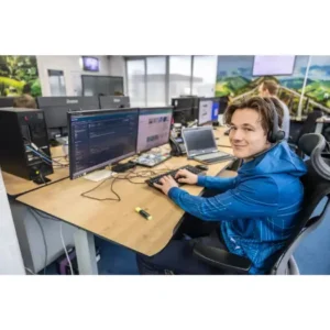 Justdata ICT, Een jonge man met een headset op en een blauwe capuchon zit aan een bureau met meerdere monitoren en typt op een toetsenbord in een moderne online werkplek met andere collega's op de achtergrond, 7 dagen per week beschikbaar.