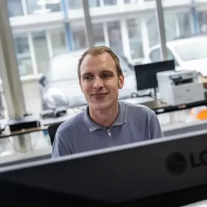Justdata ICT, Een man in een grijs overhemd met ritssluiting zit aan een bureau, glimlacht terwijl hij naar een computerscherm kijkt - misschien om automatische concepten te bekijken - in een modern kantoor met grote ramen en geparkeerde auto's zichtbaar buiten.