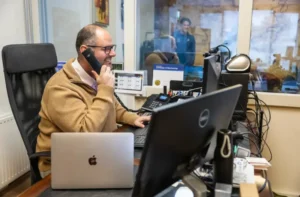 Justdata ICT, Een man met een bril en een bruine trui zit aan een bureau met meerdere computerschermen, telefoneert en glimlacht in een kantooromgeving. Op de achtergrond zijn twee mensen te zien door een glazen raam.