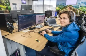 Justdata ICT, Een jongere met een headset zit aan een bureau met meerdere monitoren, typt op een toetsenbord en lacht naar de camera in een modern kantoor waar anderen op de achtergrond werken, geconcentreerd op automatische concepten.