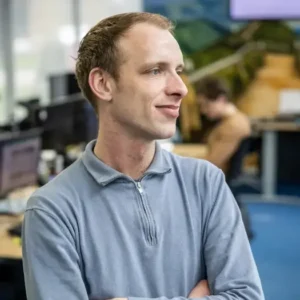 Justdata ICT, Een persoon met kort lichtbruin haar en een lichtblauwe trui met ritssluiting staat binnen met zijn armen over elkaar en glimlacht lichtjes. Bureaus, computers en een andere persoon zijn zichtbaar in de onscherpe achtergrond.