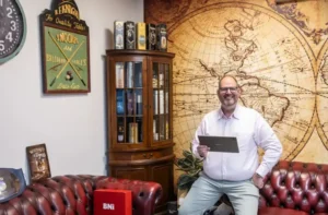 Justdata ICT, Een lachende man in een wit overhemd zit op een roodleren bank en houdt een tablet vast. Achter hem staan vintage decorstukken, een houten kast en een grote muurschildering van een landkaart. De omgeving lijkt een kantoor of studeerkamer te zijn.