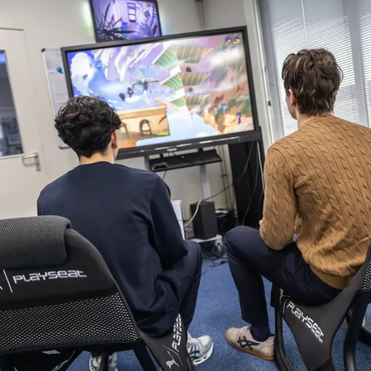 Justdata ICT, Twee mensen zitten op Playseat-gamesstoelen en spelen een videogame op een groot tv-scherm in een lichte kamer. De actie is kleurrijk, maar af en toe is er een vertraging die wijst op wifi-problemen - een van de vele wifi-oorzaken in gamingopstellingen.