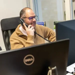 Justdata ICT, Een man met een bril en een bruine trui zit aan een bureau, glimlacht terwijl hij praat door een telefoon met snoer, met computerschermen voor hem in een kantooromgeving.