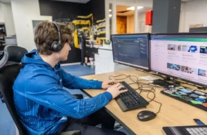 Justdata ICT, Een persoon met een koptelefoon op en een blauwe capuchon typt op een toetsenbord aan een bureau met twee grote monitoren, waarvan de ene een code weergeeft en de andere een website. Kantoorapparatuur en muurdecoraties zijn zichtbaar op de achtergrond.