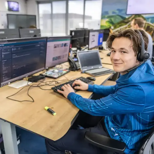 Justdata ICT, Een jonge man met een headset zit aan een bureau met meerdere monitoren en een laptop, typt op een toetsenbord en lacht naar de camera in een moderne online werkplek kantooromgeving.