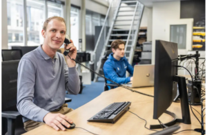 Justdata ICT, Twee mensen werken in een modern kantoor. Eén persoon zit aan een bureau en lacht terwijl hij praat via een telefoon met snoer en een computer gebruikt. Een andere persoon typt op een laptop op de achtergrond bij een trap.