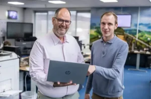 Justdata ICT, Twee mannen staan in een modern kantoor, glimlachen en houden samen een open HP-laptop vast, met bureaus, computers en grote ramen op de achtergrond.
