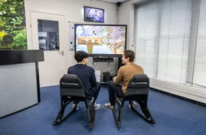 Justdata ICT, Twee mensen zitten op zwarte gamestoelen en spelen een videospel dat wordt weergegeven op een groot scherm in een lichte kamer met blauw tapijt en luxaflex. Erboven is een kleine tv gemonteerd waarop een ander scherm te zien is.
