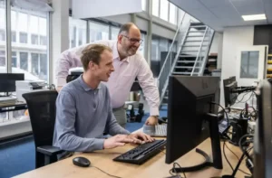 Justdata ICT, Twee mannen in een slank ingericht kantoor werken samen achter een computer en gebruiken Microsoft 365. De een zit, typt en glimlacht, terwijl de ander naast hem staat. De moderne werkruimte heeft grote ramen en bureaus, waardoor een lichte en veilige omgeving ontstaat.