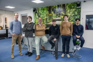 Justdata ICT, Zes mannen poseren en glimlachen in een kantoor met blauw tapijt en een grote muurschildering van een weelderig groen bos en een houten brug achter hen; sommigen staan, anderen zitten op een kruk.