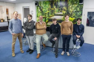 Justdata ICT, Zes mensen poseren binnenshuis voor een grote muurschildering van een bos en een houten brug; vijf zitten op barkrukken en één staat, allemaal gekleed in vrijetijdskleding.