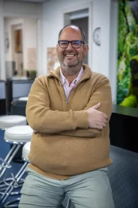 Justdata ICT, Een lachende man met bril en baard, die een bruine trui en een lichtgrijze broek draagt, zit op een stoel met zijn armen over elkaar in een moderne kantoorruimte.