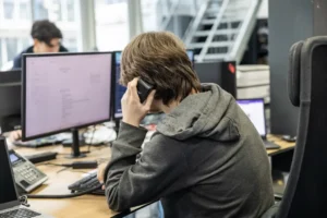 Justdata ICT, Een persoon met een grijze capuchon zit aan een bureau met meerdere computerschermen, typt op een toetsenbord en telefoneert in een modern kantoor. Een andere persoon werkt op de achtergrond.