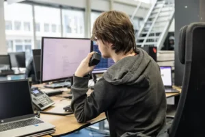 Justdata ICT, Een persoon met schouderlang haar en een capuchon zit aan een bureau in een kantoor, kijkt naar een computerscherm en spreekt in een handheld radio. Op het bureau staan meerdere monitoren, een laptop en een telefoon.