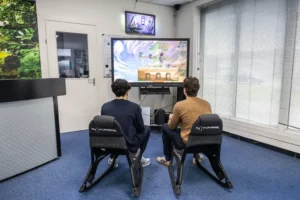 Justdata ICT, Twee mensen zitten op gamestoelen voor een grote tv en spelen een videogame in een lichte kamer met blauw tapijt, grote ramen en een moderne inrichting. Kabels en controllers zijn zichtbaar bij hun voeten.
