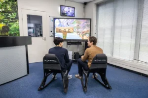 Justdata ICT, Twee mensen zitten in gamestoelen en spelen een videogame op een groot scherm in een lichte kamer met blauw tapijt, luxaflex en een kleine tv boven de deur.