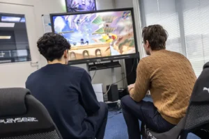 Justdata ICT, Twee mensen zitten in gamestoelen tegenover een grote monitor en spelen samen een kleurrijk vechtspel in een lichte kamer met ramen en jaloezieën.