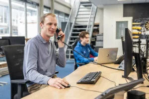 Justdata ICT, Twee mannen zitten aan een bureau in een modern kantoor. De man op de voorgrond lacht en praat met een bureautelefoon terwijl hij een computer gebruikt. De man op de achtergrond typt op een laptop.