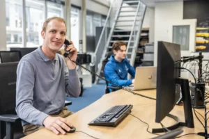 Justdata ICT, Twee mensen werken in een modern kantoor; de ene persoon zit aan een bureau en gebruikt een computer en praat met een bedrade telefoon terwijl hij lacht, de andere werkt op een laptop op de achtergrond.