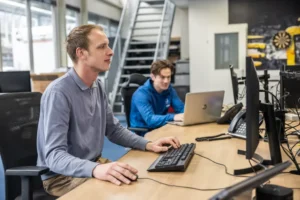 Justdata ICT, Twee mannen werken aan bureaus in een modern kantoor; één typt op een desktop computer op de voorgrond terwijl de ander een laptop gebruikt op de achtergrond. Er zijn stoelen, telefoons en grote ramen.