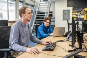 Justdata ICT, Twee mensen werken achter een computer in een modern kantoor. Eén persoon zit op de voorgrond te typen op een toetsenbord, terwijl de ander op de achtergrond een laptop gebruikt. Kantoorapparatuur en een trap zijn zichtbaar.