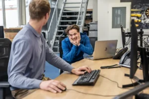 Justdata ICT, Twee mannen zitten aan een bureau met computers in een kantoor. De ene man in het blauw leunt voorover, glimlacht met zijn kin in zijn handen, terwijl de andere zich naar hem toe draait, met één hand op het toetsenbord. Op de achtergrond zijn trappen en planken te zien.