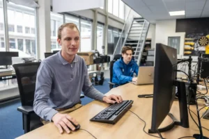 Justdata ICT, Twee mensen werken aan bureaus in een modern kantoor. De ene persoon gebruikt een desktop computer op de voorgrond, terwijl de andere op een laptop werkt op de achtergrond. Grote ramen en planken zijn zichtbaar.