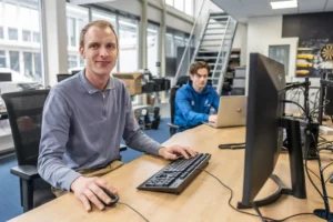 Justdata ICT, Twee mannen werken achter een computer in een modern kantoor met grote ramen. De ene man in een grijs shirt kijkt en glimlacht naar de camera, terwijl de andere, in het blauw, zich concentreert op zijn laptop. Kantoorapparatuur en een trap zijn zichtbaar.