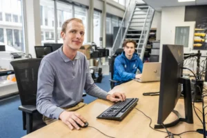 Justdata ICT, Twee mensen werken achter een computer in een licht, modern kantoor. Een persoon in een grijs shirt zit aan een bureau op de voorgrond en kijkt naar de camera, terwijl een ander in blauw op de achtergrond aan een laptop werkt.