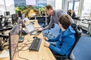 Justdata ICT, Twee mensen in een kantoor kijken naar een laptop; de een zit aan een bureau met computerschermen, terwijl de ander staat en naar het scherm wijst, wat een gezamenlijke werkbespreking suggereert.