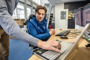 Justdata ICT, Een jonge man in een blauw jasje zit aan een bureau met computers en kijkt toe terwijl een andere persoon iets aanwijst op het toetsenbord van een laptop. De setting lijkt een modern kantoor met grote ramen en een trap op de achtergrond.