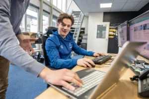 Justdata ICT, Twee mensen werken samen in een modern kantoor; de een zit aan een bureau met twee beeldschermen en een toetsenbord, terwijl de ander vlakbij staat en op een laptop wijst. Op de achtergrond zijn dozen en kantoorapparatuur zichtbaar.