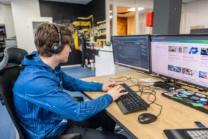 Justdata ICT, Een persoon met een koptelefoon op en een blauwe capuchon zit aan een bureau met een toetsenbord en muis en werkt op twee grote computermonitoren die code en webinhoud weergeven in een modern kantoor.