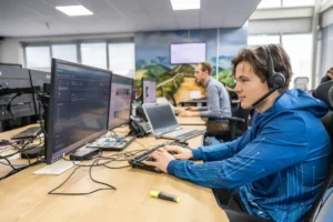 Justdata ICT, Een persoon met een headset werkt aan een computer in een modern kantoor, met een andere persoon aan een bureau op de achtergrond. Meerdere monitoren, laptops en kantoorbenodigdheden zijn zichtbaar op de bureaus.