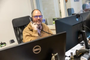 Justdata ICT, Een man met een bril en een bruine trui zit aan een bureau in een kantoor en praat met een telefoon met snoer. Hij kijkt naar twee computerschermen en op het bureau zijn kantoorbenodigdheden en papieren zichtbaar.