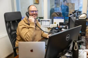 Justdata ICT, Een lachende man met bril en bruine trui zit aan een bureau met een telefoon aan zijn oor, omringd door een laptop, computermonitor en kantoorbenodigdheden in een helder, modern kantoor.