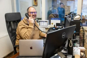 Justdata ICT, Een man met een bril en een bruine trui zit aan een bureau en lacht terwijl hij telefoneert. Hij wordt omringd door computerschermen, een laptop en kantoorbenodigdheden. Een andere persoon is zichtbaar door een raam achter hem.