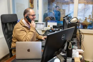 Justdata ICT, Een man met een bril en een bruine trui zit aan een bureau met meerdere computers en een telefoon en lacht terwijl hij telefoneert in een kantooromgeving. Een andere persoon is zichtbaar door een raam op de achtergrond.