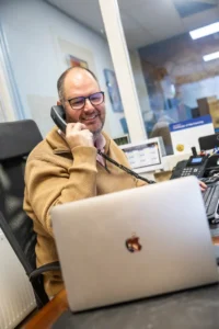 Justdata ICT, Een lachende man met een bril en een bruine trui zit aan een bureau te praten met een telefoon met snoer. Hij kijkt uit op een open laptop en heeft verschillende kantoorartikelen om zich heen, werkend in een moderne kantooromgeving.