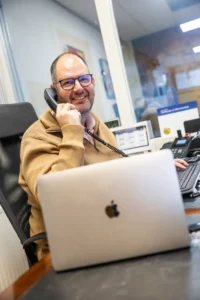 Justdata ICT, Een man met een bril en een bruine trui lacht terwijl hij praat aan een bedrade telefoon aan zijn bureau, met een open laptop, een toetsenbord en kantoorapparatuur voor hem.