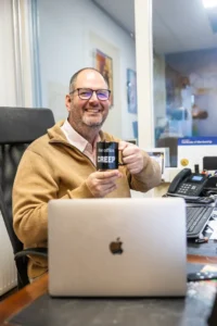 Justdata ICT, Een lachende man met een bril en een bruine trui zit aan een bureau met een laptop en houdt een zwarte mok vast waarop CREEP staat. Kantoorbenodigdheden, een telefoon en papieren zijn zichtbaar op het bureau.
