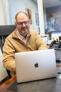 Justdata ICT, Een man met een bril en een bruine trui zit aan een bureau en werkt op een zilveren MacBook laptop. Kantoorbenodigdheden, een telefoon en documenten zijn zichtbaar op het bureau. Hij lijkt geconcentreerd en tevreden.