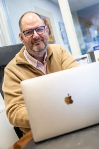 Justdata ICT, Een man met een bril en een baard, die een bruine trui draagt over een overhemd met col, zit aan een bureau en glimlacht terwijl hij werkt op een zilverkleurige Apple laptop in een lichte kantooromgeving.