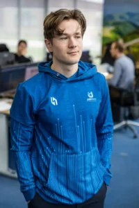 Justdata ICT, Een jong persoon met lichtbruin haar draagt een blauwe hoodie met tech-thema patronen, staand in een kantoor met vage mensen en bureaus op de achtergrond.