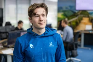 Justdata ICT, Een jonge man met een blauwe capuchon staat binnen in een kantooromgeving, met andere mensen aan bureaus op de achtergrond. De sfeer is casual en modern.