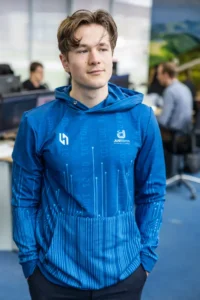 Justdata ICT, Een jonge man met lichtbruin haar die een blauwe hoodie draagt met witte logo's en binaire codepatronen staat binnen, met bureaus en mensen aan het werk op de achtergrond.