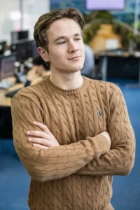 Justdata ICT, Een jonge man met lichtbruin haar en een bruine kabeltrui staat binnen met zijn armen over elkaar en kijkt opzij. Op de achtergrond zijn bureaus en computers te zien.