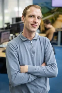 Justdata ICT, Een man in een lichtblauwe trui staat binnen met zijn armen over elkaar en glimlacht zelfverzekerd. Bureaus, computers en een andere persoon die op de achtergrond aan het werk is, zijn zichtbaar.